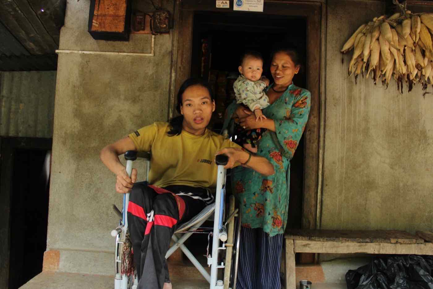 Renuka Tamang holding her daughter beside her husband, who uses a wheelchair, outside their home and shop in Kamchok