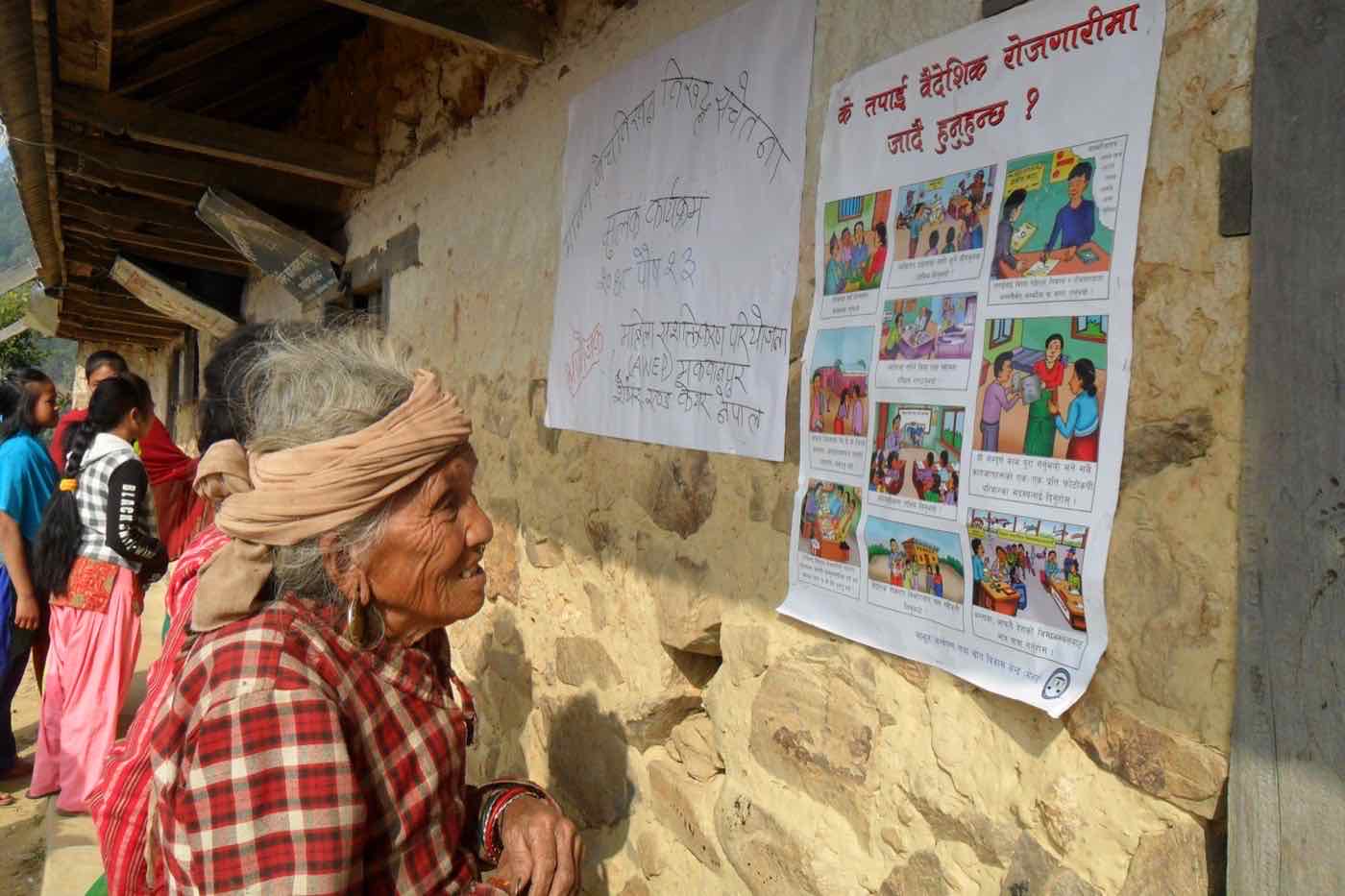 An elderly woman reading AWEP safe-migration posters on a village wall in Makwanpur