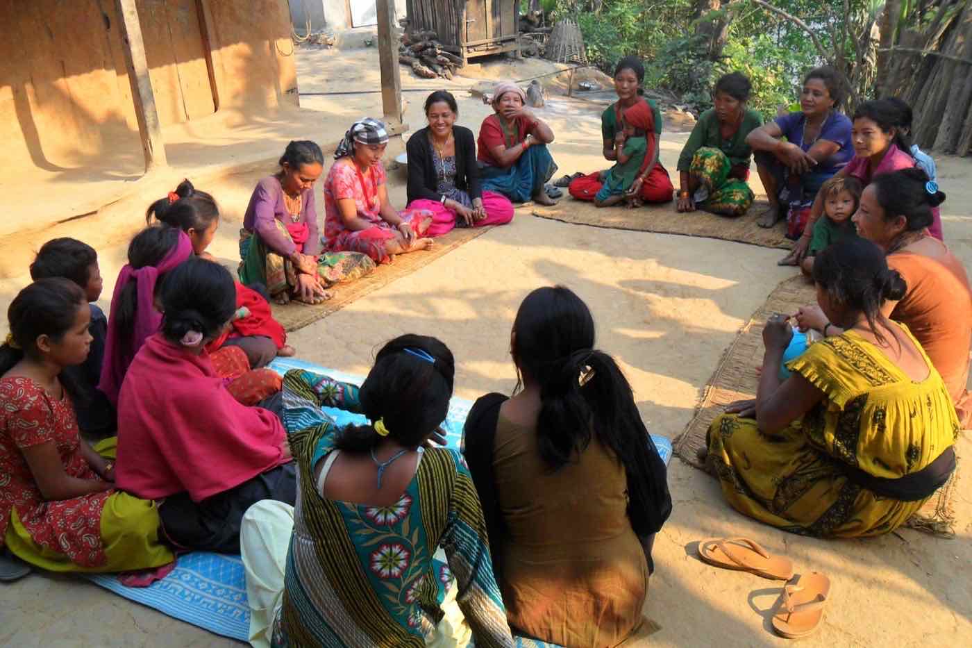 Smiling women seated in a circle on mats in a sunlit village courtyard