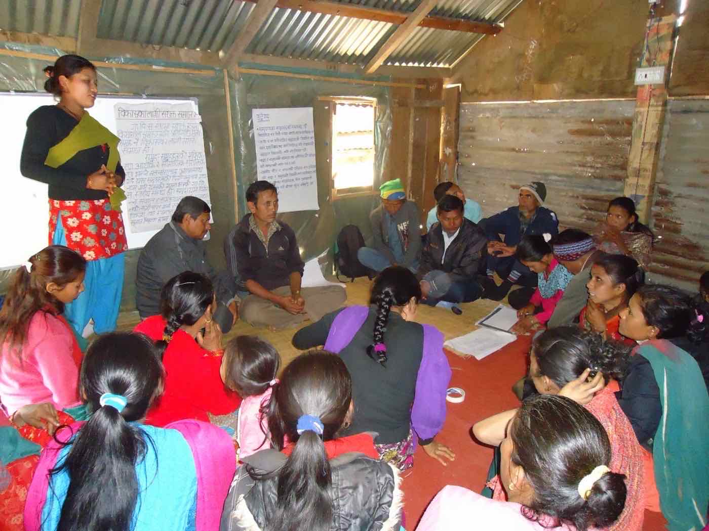 A woman leading a discussion with men and women seated inside a tin-roofed meeting hall