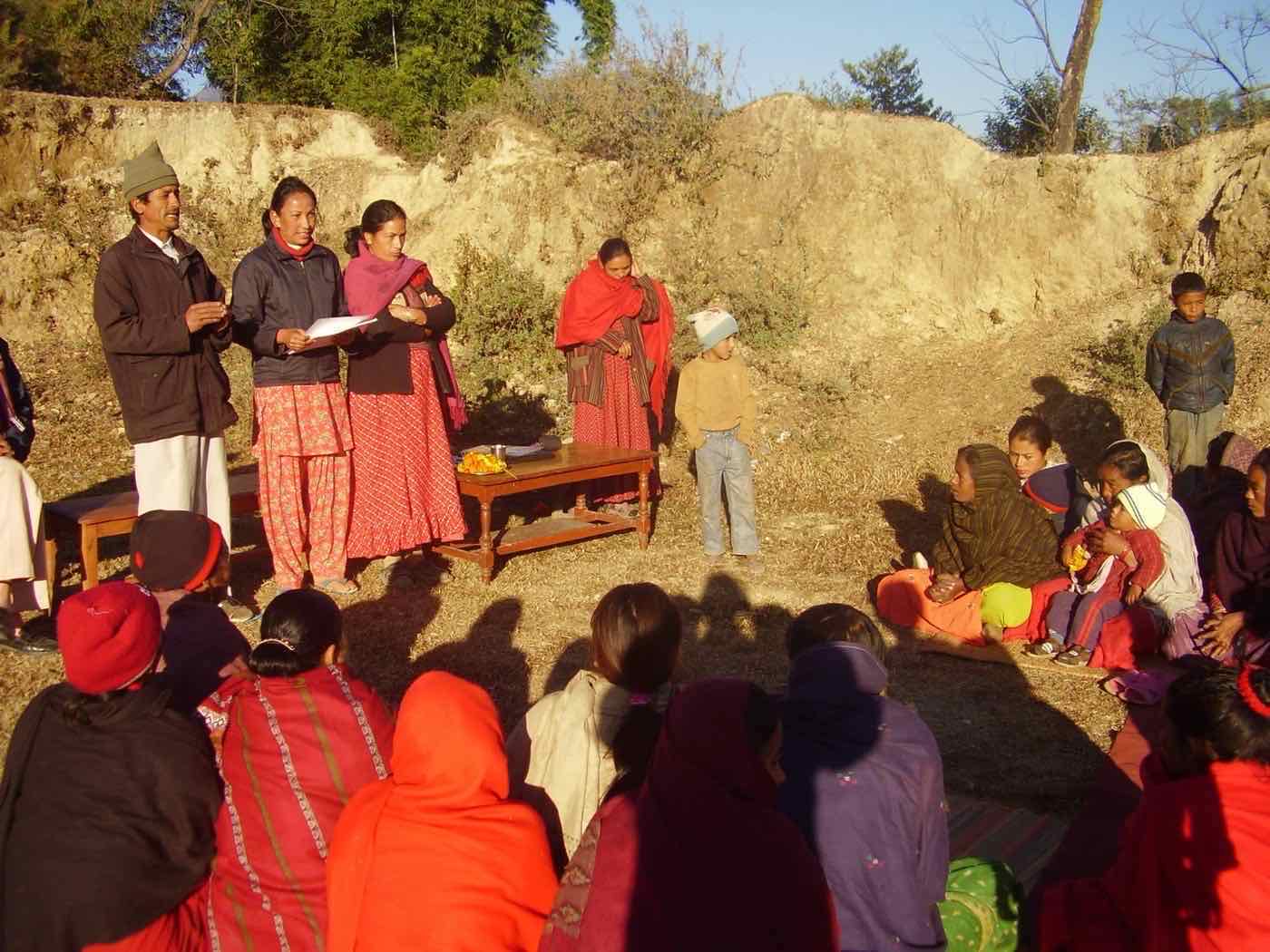 Facilitators addressing villagers seated in a circle at an outdoor meeting