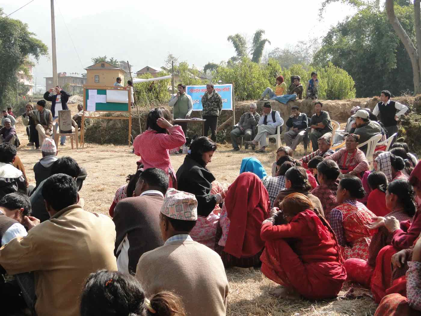 A community meeting seated outdoors around a speaker and a water user committee banner, Empowering Through Safe Water and Agriculture (ETSWA) project in Lamatar, Lalitpur